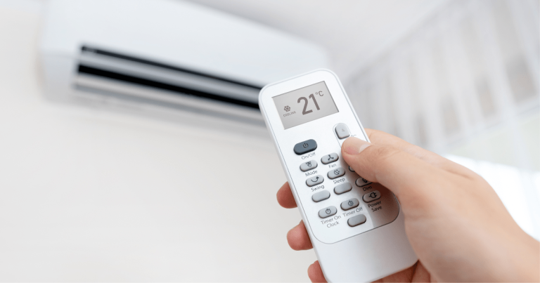 Hand holding a remote control set to 21°C in front of a wall-mounted air conditioner unit.